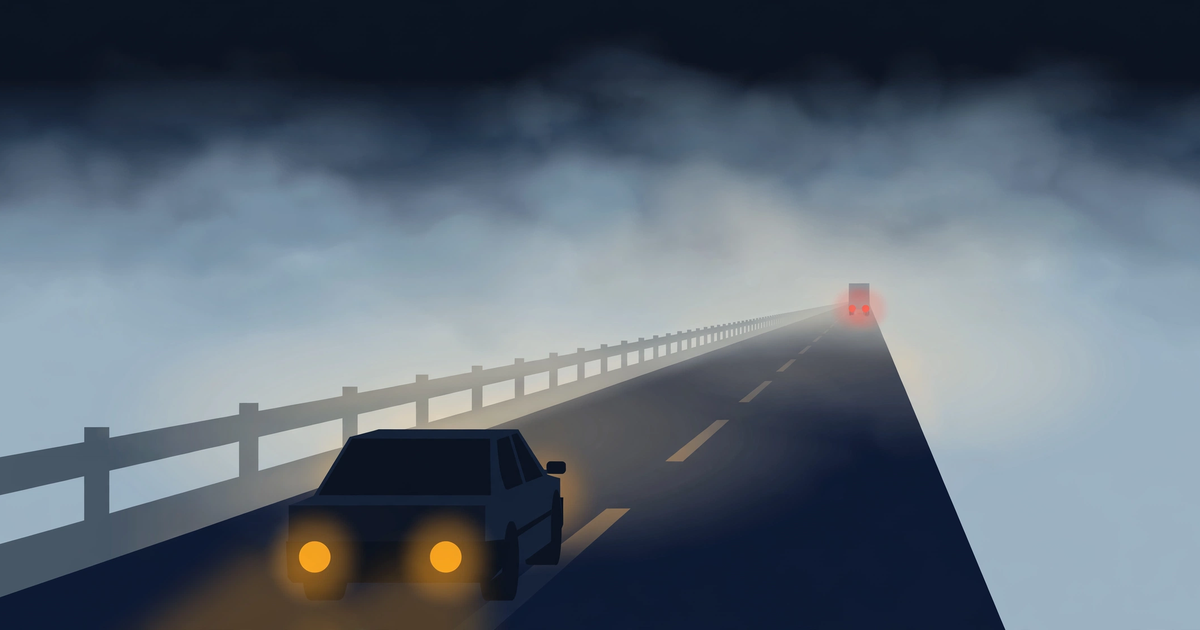 A car with low amber fog lights on a dark motorway disappearing into thick fog, with the faint red tail lights of a truck far ahead and a guardrail fading into white